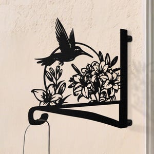 Könnte beinhalten: Schwarze Metall-Wandkunst mit einem Kolibri und Blumen. Das Design zeigt einen Kolibri im Flug über einer Ansammlung von Blumen und Blättern. Das Kunstwerk ist zum Aufhängen an der Wand konzipiert.