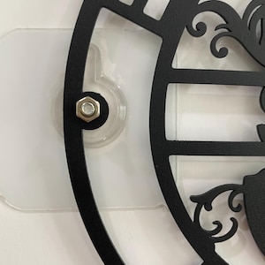 May include: Close-up of a black metal decorative wall clock with a cut-out design. The clock is attached to a clear plastic adhesive mount with a silver bolt. The clock's design includes ornate scrollwork and a partial view of the clock face.