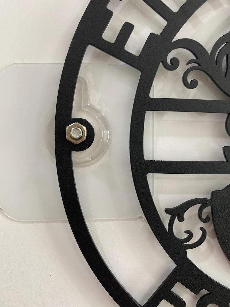May include: Close-up of a black metal decorative wall clock with a cut-out design. The clock is attached to a clear plastic adhesive mount with a silver bolt. The clock's design includes ornate scrollwork and a partial view of the clock face.