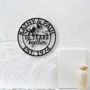 May include: Black metal wall art with the names "KATHY & PAUL" encircling the design. The art features two birds perched on a branch, with the text "50 YEARS Together EST. 1974" in the center. The art is mounted on a white wall.