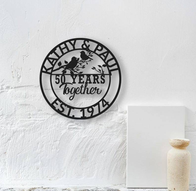 May include: Black metal wall art with the names "KATHY & PAUL" encircling the design. The art features two birds perched on a branch, with the text "50 YEARS Together EST. 1974" in the center. The art is mounted on a white wall.