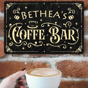 May include: A black metal sign with the text "BETHEA'S COFFE BAR" in a decorative font. The sign has a distressed look with coffee bean and cup illustrations. The sign is hanging on a brick wall.