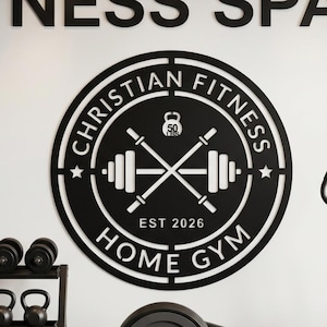 Könnte beinhalten: Schwarz-weißes Metallschild mit den Worten "CHRISTIAN FITNESS HOME GYM" in einem kreisförmigen Design. Das Schild enthält gekreuzte Langhanteln, ein Gewicht und den Text "EST 2026". Es ist an einer weißen Wand befestigt.