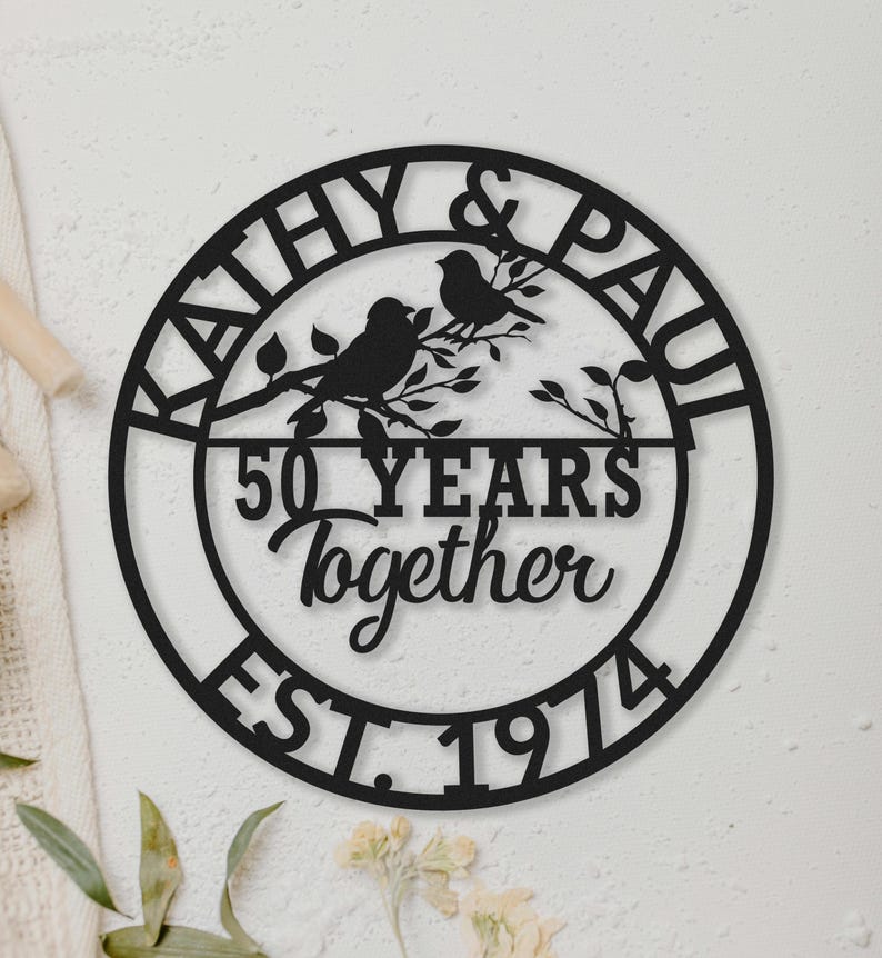 May include: Black metal wall art with the names "KATHY & PAUL" and the text "50 YEARS Together EST. 1974" in a circular design. Two birds perch on a branch in the center. A decorative piece for a wedding anniversary.