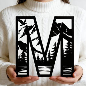 Peut inclure: Lettre "M" en bois noir avec une scène de montagne et de forêt découpée. Le design comprend un aigle, des arbres, des montagnes et un lac. La lettre est tenue devant un pull blanc.
