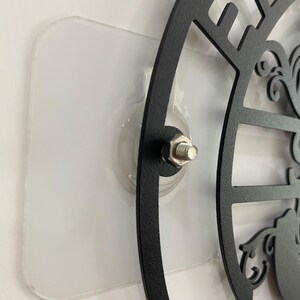 May include: Black metal wall art with a decorative design, mounted on a clear adhesive pad. The art features cut-out shapes and a central design element, attached with a bolt.