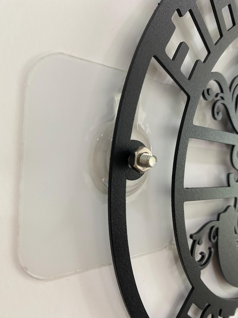 May include: Black metal wall art with a decorative design, mounted on a clear adhesive pad. The art features cut-out shapes and a central design element, attached with a bolt.