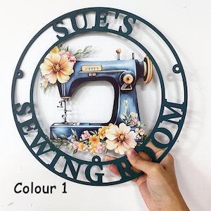 Puede incluir: Un letrero redondo de color verde oscuro con las palabras "SUE'S SEWING ROOM" recortadas. Dentro del círculo hay una máquina de coser azul con detalles florales. El texto "Colour 1" está en la parte inferior.