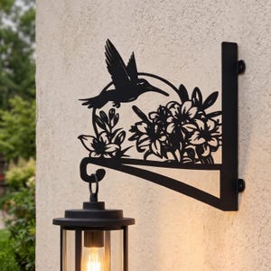 May include: A black metal wall-mounted bracket featuring a silhouette of a hummingbird hovering over a cluster of flowers. The bracket includes a curved hook at the bottom designed to support a hanging lantern, which is shown illuminated below.