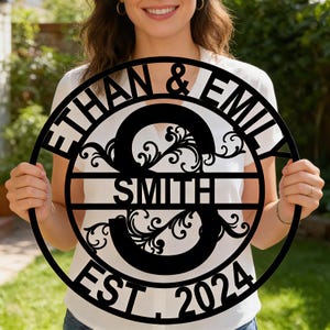 May include: A black metal family name sign with the names "ETHAN & EMIL" at the top, "SMITH" in the center, and "EST. 2024" at the bottom. The sign features a large, stylized "S" with decorative flourishes.