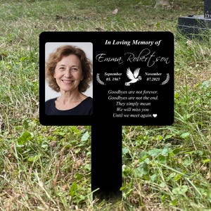 Pode incluir: Placa memorial preta com foto de mulher, texto "In Loving Memory of Emma Robertson" e datas. A placa inclui o texto "Goodbyes are not forever. We will miss you."