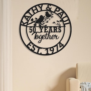 May include: Black metal wall art with the names "Kathy & Paul" and the text "50 YEARS Together EST. 1974". The art features two birds perched on a branch within a circular frame, suitable for a wedding or anniversary gift.