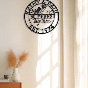 May include: Black metal wall art with the names "Kathy & Paul" and the text "50 YEARS Together EST. 1974" in a circular design. Two birds perch on a branch. The art is mounted on a white wall.