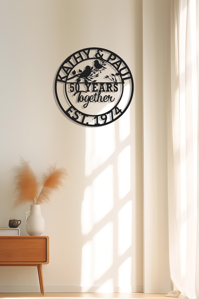 May include: Black metal wall art with the names "Kathy & Paul" and the text "50 YEARS Together EST. 1974" in a circular design. Two birds perch on a branch. The art is mounted on a white wall.