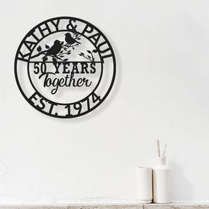 May include: Black metal wall art with a circular design. The art piece features the names "Kathy & Paul" around the top, with "50 YEARS together EST. 1974" in the center. Two birds perch on a branch. Decorative home decor.