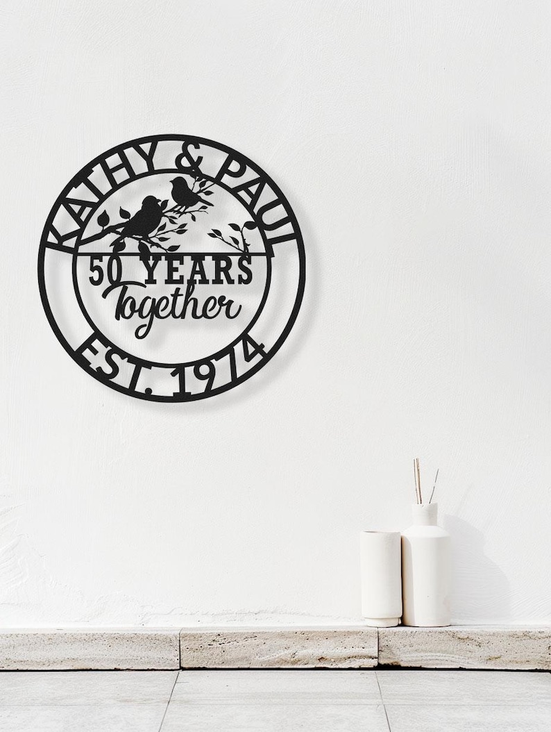 May include: Black metal wall art with a circular design. The art piece features the names "Kathy & Paul" around the top, with "50 YEARS together EST. 1974" in the center. Two birds perch on a branch. Decorative home decor.
