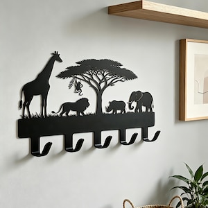 May include: Black metal wall-mounted coat rack depicting a savanna scene with a giraffe, lion, rhino, and elephant. The design includes a tree and six hooks for hanging items. The rack is mounted on a white wall.
