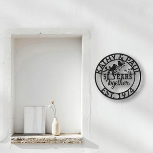 May include: Black metal wall art with the names "Kathy & Paul", two birds on a branch, and the text "50 years together EST. 1974". The art is circular and mounted on a white wall.