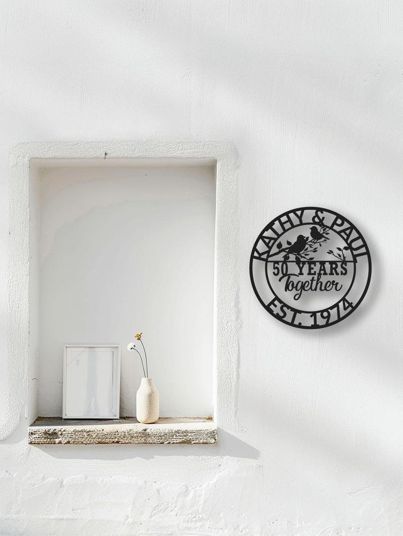 May include: Black metal wall art with the names "Kathy & Paul", two birds on a branch, and the text "50 years together EST. 1974". The art is circular and mounted on a white wall.