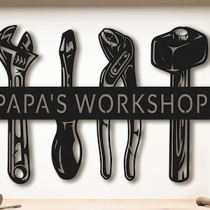 May include: Black metal sign with workshop tools: an adjustable spanner, screwdriver, pliers, and a mallet. The sign reads "PAPA'S WORKSHOP" in a bold font, ideal for a garage or workshop. A decorative piece.