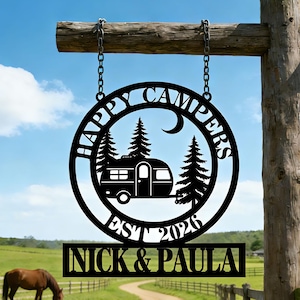 May include: Black metal sign with the words "HAPPY CAMPERS" in a circular design, featuring a camper, trees, and a crescent moon. The sign also includes "EST 2026" and the names "NICK & PAULA". The sign is suspended from a wooden beam.