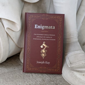 May include: A maroon book titled "Enigmata" with gold lettering and decorative borders. The book's cover includes the text "One Hundred Lyrical Riddles" and the author's name, Joseph Kay. The book is resting on a white surface.