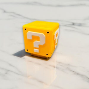 May include: A yellow cube-shaped object with white question marks on two sides. The object has a small base and black dots on the corners. The background is a white marble surface. This item is reminiscent of a video game.