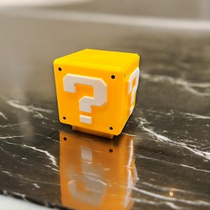 May include: A yellow cube with a white question mark on each side. The cube has black dots on the corners and is sitting on a reflective surface. This item is likely a decorative piece or a toy.