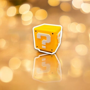 May include: A yellow cube with a white outline and a large white question mark on the front. The cube has a reflective surface and is likely a toy or decorative item. The background is blurred with golden bokeh.