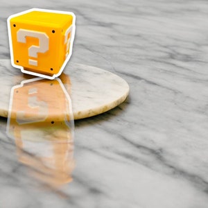 May include: A yellow cube with white question marks on each side, reminiscent of a video game item. The cube is set on a white and tan marble-like surface.