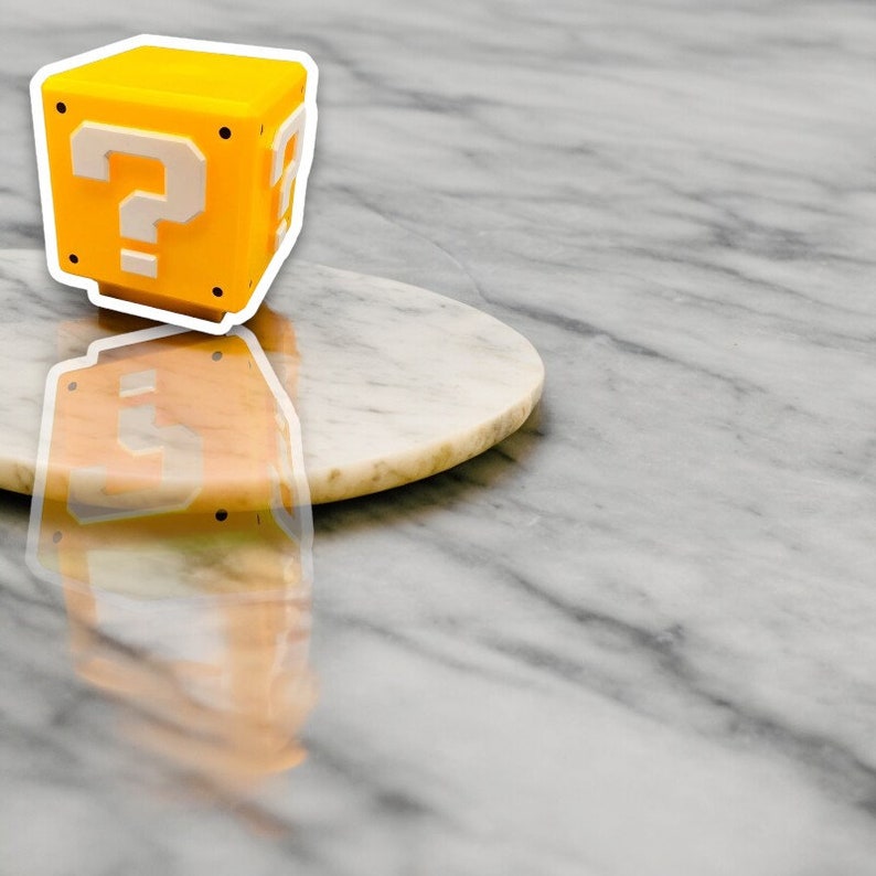 May include: A yellow cube with white question marks on each side, reminiscent of a video game item. The cube is set on a white and tan marble-like surface.