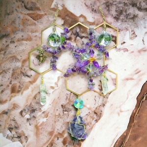 Puede incluir: Adorno colgante decorativo con una estructura dorada de panal, adornado con virutas de amatista morada, cristales de cuarzo transparente y un pequeño dije de abeja. También incluye una gran piedra de amatista envuelta en alambre y un cristal facetado.