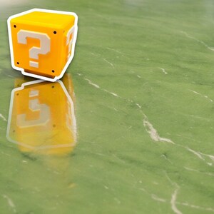 May include: A yellow cube with a white question mark on each side, reminiscent of a video game mystery block. The cube is set against a green background, with its reflection visible below.