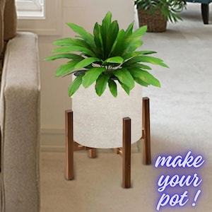 Op de afbeelding: Een grote groene plant in een witte cilindrische pot op een houten plantenstandaard. De standaard heeft vier poten en is gemaakt van donkerbruin hout. De tekst "make your pot!" staat rechtsonder.