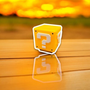May include: A yellow cube with a white outline and a white question mark on the front. The cube is reflected on a wooden surface. The background is a blurred sunset.