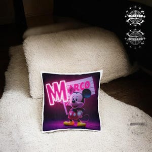 Puede incluir: Cojín decorativo cuadrado con Mickey Mouse sosteniendo un letrero que dice "Marco" en letras de neón rosa. El cojín tiene un fondo negro con un brillo púrpura, y Mickey lleva pantalones cortos rojos y zapatos amarillos.
