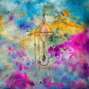 Puede incluir: Un atrapasoles geométrico dorado con un cristal rosa en el centro y cuentas de cristal colgantes. El atrapasoles está suspendido de una cadena con un diseño de estrella en la parte superior. El fondo es una pintura abstracta colorida.