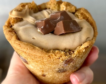 May include: A close-up of a cookie cup filled with a light brown cream and topped with chocolate pieces. The cookie cup has a golden-brown color with visible chocolate chips. The cream filling has a smooth texture.