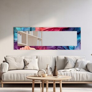 May include: A long rectangular mirror with a colorful abstract design frame. The frame features swirls of pink, blue, and purple. The mirror is hanging on a white wall above a light gray couch.