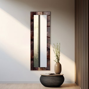 May include: A tall, rectangular mirror with a brown, textured frame. The mirror is hanging on a white wall. A small, round table with a black, woven top sits in front of the mirror. A vase with green stems and white flowers sits on the table.