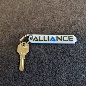 May include: A white keychain with a blue border and the word "ALLIANCE" in black lettering. A silver key is attached to the keychain.