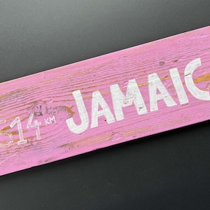 Könnte beinhalten: Ein rosafarbenes Holzschild mit weißer Beschriftung, auf dem "Jamaica" und die Zahl "8,14" mit den Buchstaben "KM" daneben stehen.