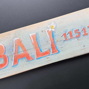Könnte beinhalten: Ein Holzschild mit dem Wort "BALI" in orangefarbenen Buchstaben mit blauen Konturen. Die Zahl "11513 km" ist ebenfalls orange. Auf dem hellblauen Schild sind weiße Blumen gemalt. Das Schild hat ein rustikales Aussehen.
