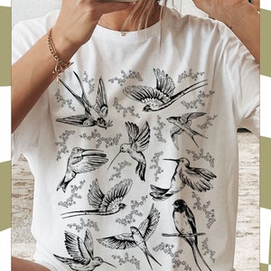 K&ouml;nnte beinhalten: Wei&szlig;es T-Shirt mit einem schwarz-wei&szlig;en grafischen Druck von V&ouml;geln und floralen Designs. Das Shirt hat einen Rundhalsausschnitt und kurze &Auml;rmel.
