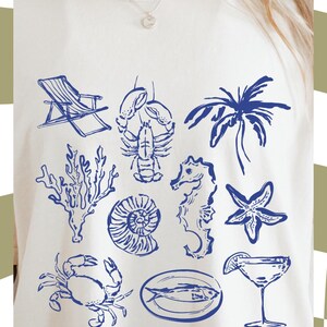 Op de afbeelding: Cr&egrave;mekleurig T-shirt met blauwe nautische illustraties, waaronder een strandstoel, kreeft, palmboom, koraal, zeepaardje, zeester, krab, schelp, vis en een cocktail. Tekst: "GRATIS COMMERCIEEL GEBRUIK!" en "PNG FILE".