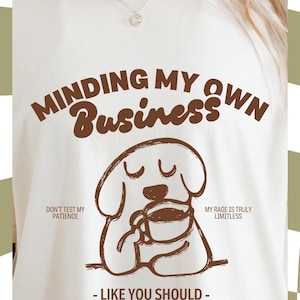 May include: White t-shirt with the phrase "MINDING MY OWN BUSINESS" in brown text. A cartoon dog illustration holding a coffee cup is below the text. Additional text includes "DON'T TEST MY PATIENCE" and "MY RAGE IS TRULY LIMITLESS".