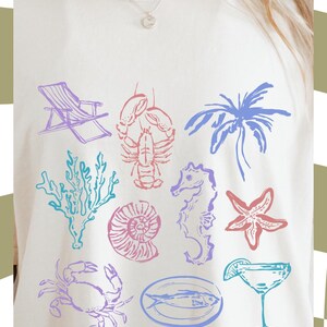 May include: A white t-shirt featuring a colourful design of various sea creatures, including a lobster, a seahorse, a starfish, a seashell, a crab, a fish, a palm tree, and a deckchair. The text "PNG FILE" is at the bottom of the shirt.