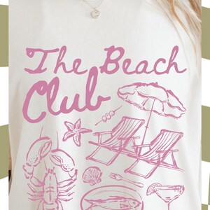 May include: A white t-shirt with a pink graphic design that says "The Beach Club". The graphic also includes a lobster, a starfish, two lounge chairs, an umbrella, a plate of fish, a cocktail glass, and a fork.
