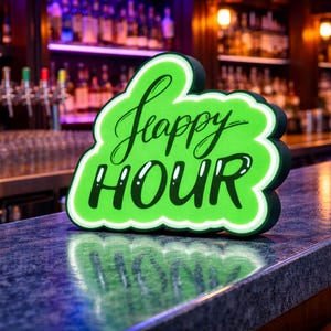 May include: A neon green "Happy Hour" sign with a white border is displayed on a bar. The sign is cloud-shaped and features the words "Happy Hour" in a stylish, cursive font. The bar has a dark countertop and a blurred background.