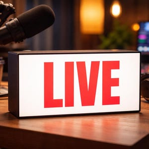 Peut inclure: Un panneau lumineux rectangulaire avec le mot "LIVE" en lettres rouges. Un microphone, un casque et un gobelet de café sont posés sur une table en bois. L'arrière-plan est flou.